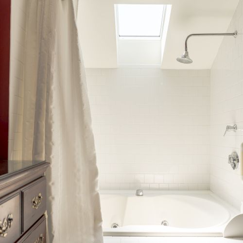 A bathroom with white tiles features a shower, bathtub, skylight, curtain, and a wooden cabinet topped with a small ceramic container.