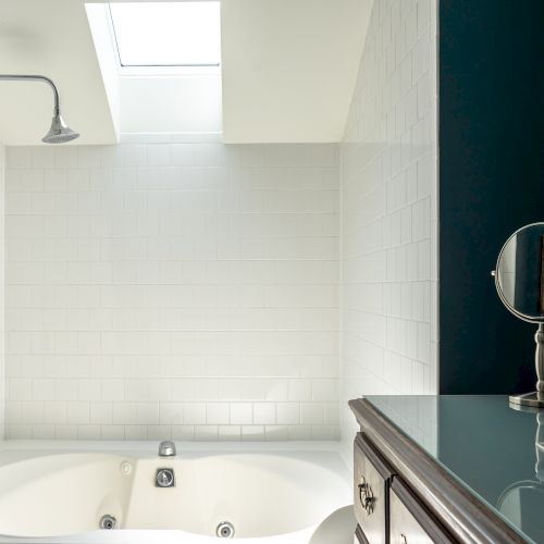 A bathroom with a bathtub, white tiled walls, a skylight above, a showerhead, a vanity with a mirror, and glass jars is shown in the image.