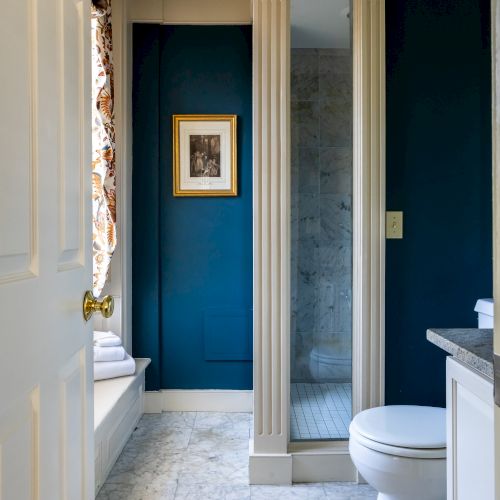 This image shows a bathroom with blue walls, a white toilet, a marble floor, a shower with a glass door, and a painting on the wall.