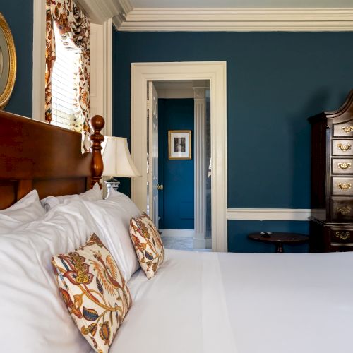 A cozy bedroom features a neatly made bed with white linens and patterned pillows, a wooden dresser, and blue walls adorned with framed artwork.