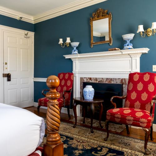 A cozy, elegant room featuring a bed, two red armchairs, a mantel with a mirror above, and blue walls, creating a warm and inviting atmosphere.