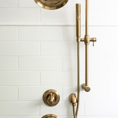 An image of a shower setup with gold-colored fixtures, including a showerhead, adjustable hand shower, and control knobs, mounted on white tiles.