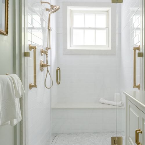 A bright, modern shower with glass doors, gold fixtures, a built-in bench, and a towel hanging outside the shower, with a small window.