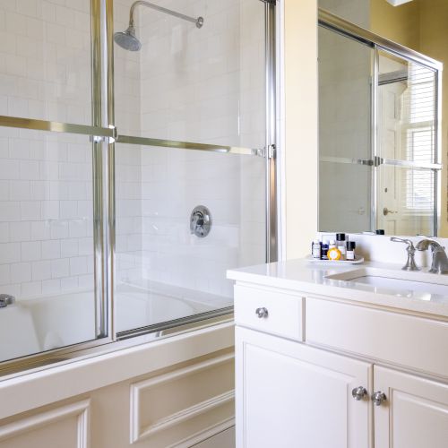 The image shows a bathroom with a glass-enclosed shower, white vanity, sink, mirror, tiled floor, and wall lights above the mirror.