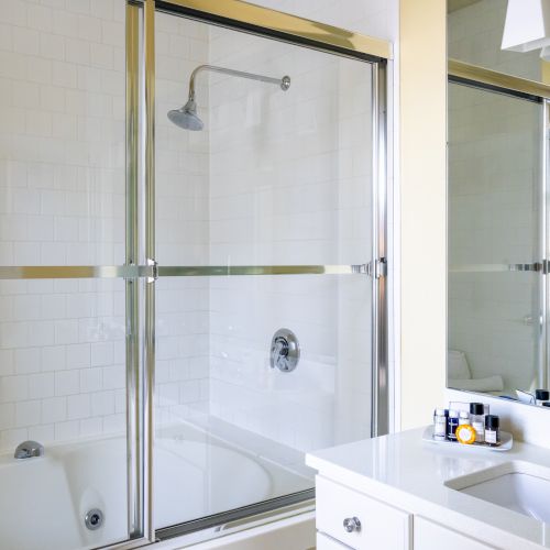 The image shows a clean bathroom with a bathtub, a glass shower door, a large mirror, a sink with toiletries, and a vanity light fixture.