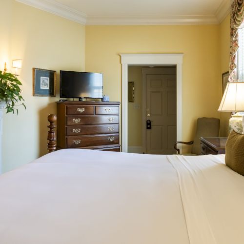 A cozy bedroom featuring a large bed with white linens, a TV on a dresser, a fireplace with a picture above, and a door leading outside.