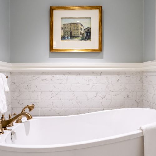 The image shows a stylish bathroom with a white freestanding bathtub, gold fixtures, a towel, and a framed picture on the wall above.