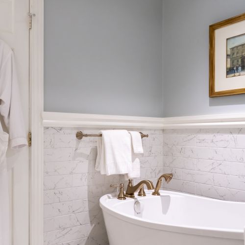 The image shows a bathroom with a white robe hanging on a door, a bathtub with gold fixtures, a towel, and a framed picture on the wall.