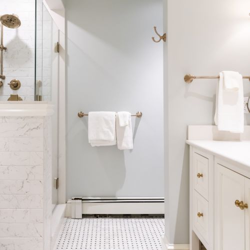 Bright bathroom with white counters, gold hardware, white shower tiles, wall-mounted towel rack, and patterned floor tiles.