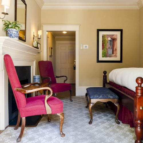 A cozy room with red chairs, a bed, fireplace, wall art, and a lamp. There's a small window and a door in the background.