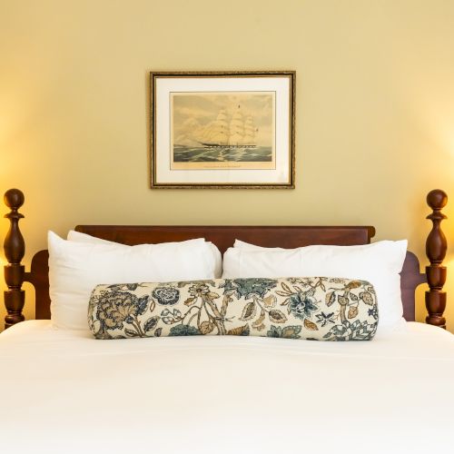 A neatly made bed with white linens, floral pillows, two bedside lamps, and a framed picture of a ship on the wall above it.