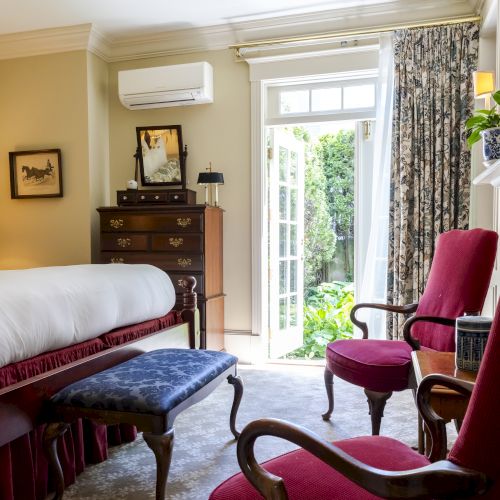 A cozy bedroom features a large bed, wooden furniture, and red chairs, with French doors opening to a garden area and natural light streaming in.