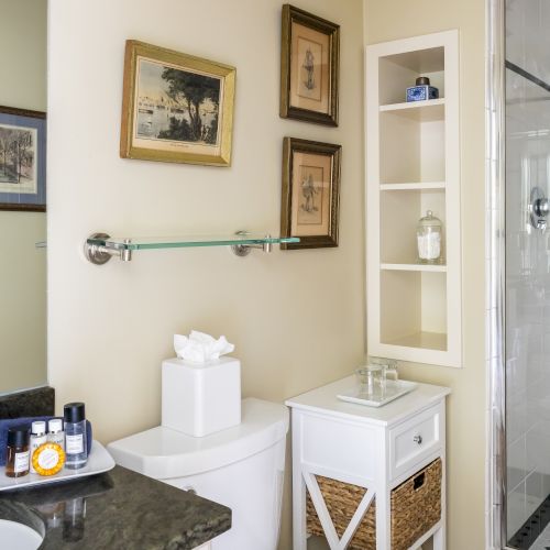 A small bathroom with a glass shower, framed pictures on the wall, a glass shelf, a tissue box, toiletries, and a white shelving unit with baskets.