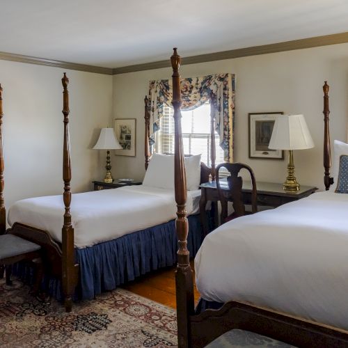 A cozy bedroom featuring two four-poster beds with white bedding, bedside tables with lamps, and a decorative rug is pictured.