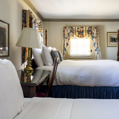 The image shows a well-lit bedroom with two neatly made beds, wooden furniture, framed artwork, floral curtains, and a bedside lamp.