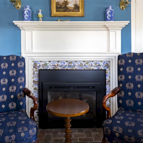 A cozy living room with blue walls features a fireplace, two blue patterned chairs, and a small round table. A painting and decor items adorn the mantel.