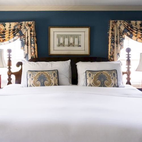 The image shows a neatly made bed with two decorative pillows, flanked by bedside tables with lamps, in a room with blue walls and floral curtains.