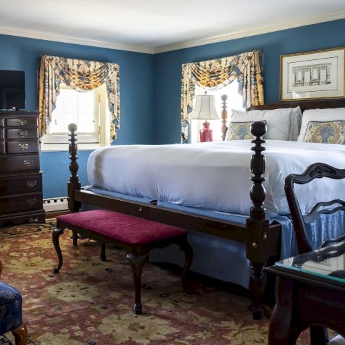 A cozy bedroom features a large wooden bed, a chest of drawers, a small bench, and traditional decor with blue walls and patterned curtains.