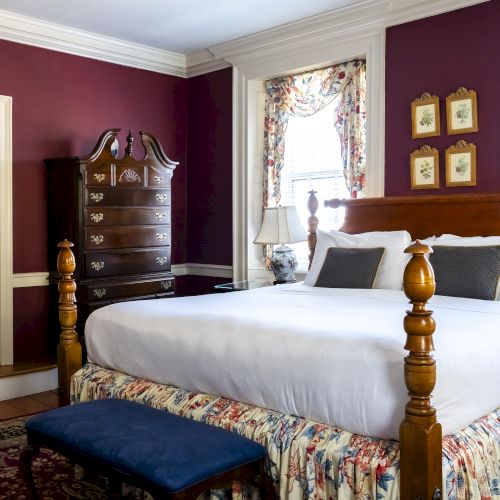 The image shows a bedroom with burgundy walls, a large bed with a floral bed skirt, a wooden dresser, two windows with floral curtains, and a door.