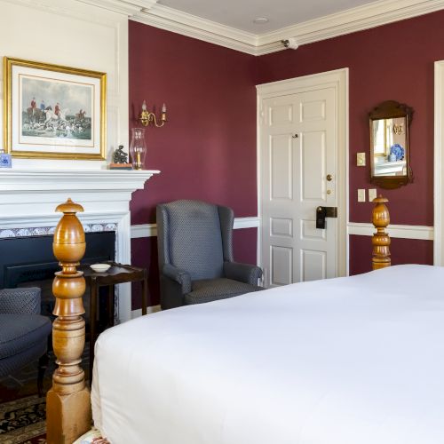 A cozy bedroom with a large bed, two armchairs, a fireplace, and maroon-colored walls, featuring charming decor and a painting above the mantel.