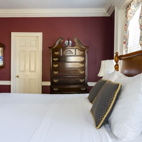 A well-decorated bedroom with a bed, pillows, a wooden dresser, framed pictures, a mirror, and doors, featuring maroon walls and white accents.