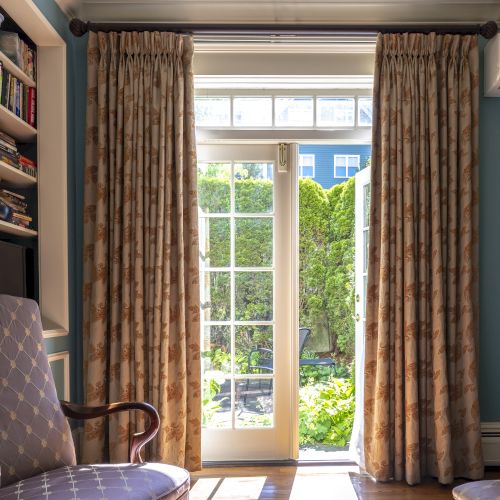 A cozy room with French doors leading to a garden, flanked by curtains, two chairs, a bookshelf, and framed artwork on a light blue wall.