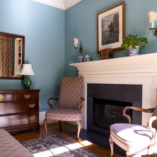 A room with blue walls, two chairs, a dresser, a mirror, a lamp, a fireplace, framed art, and decorative items is shown.