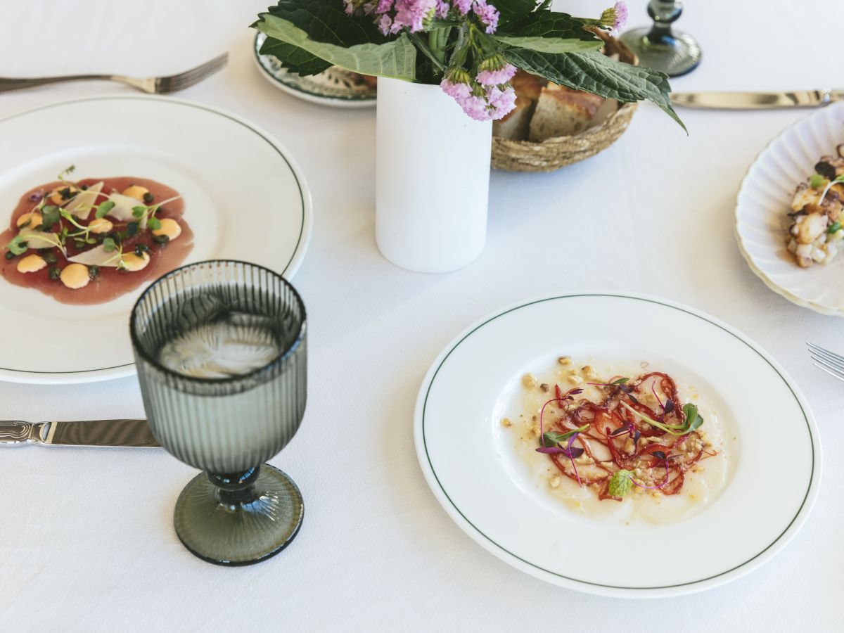 A white tablecloth with plates of gourmet food, cutlery, a green glass, and a flower vase centerpiece.