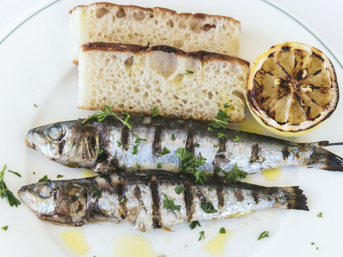 The image shows two grilled fish garnished with herbs, accompanied by slices of bread and a grilled lemon half on a white plate.