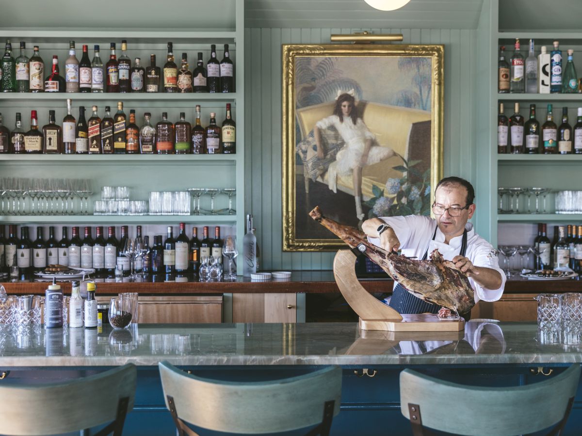 A bartender prepares a dish behind a marble bar in a well-stocked bar with a portrait of a woman and dog on the wall.