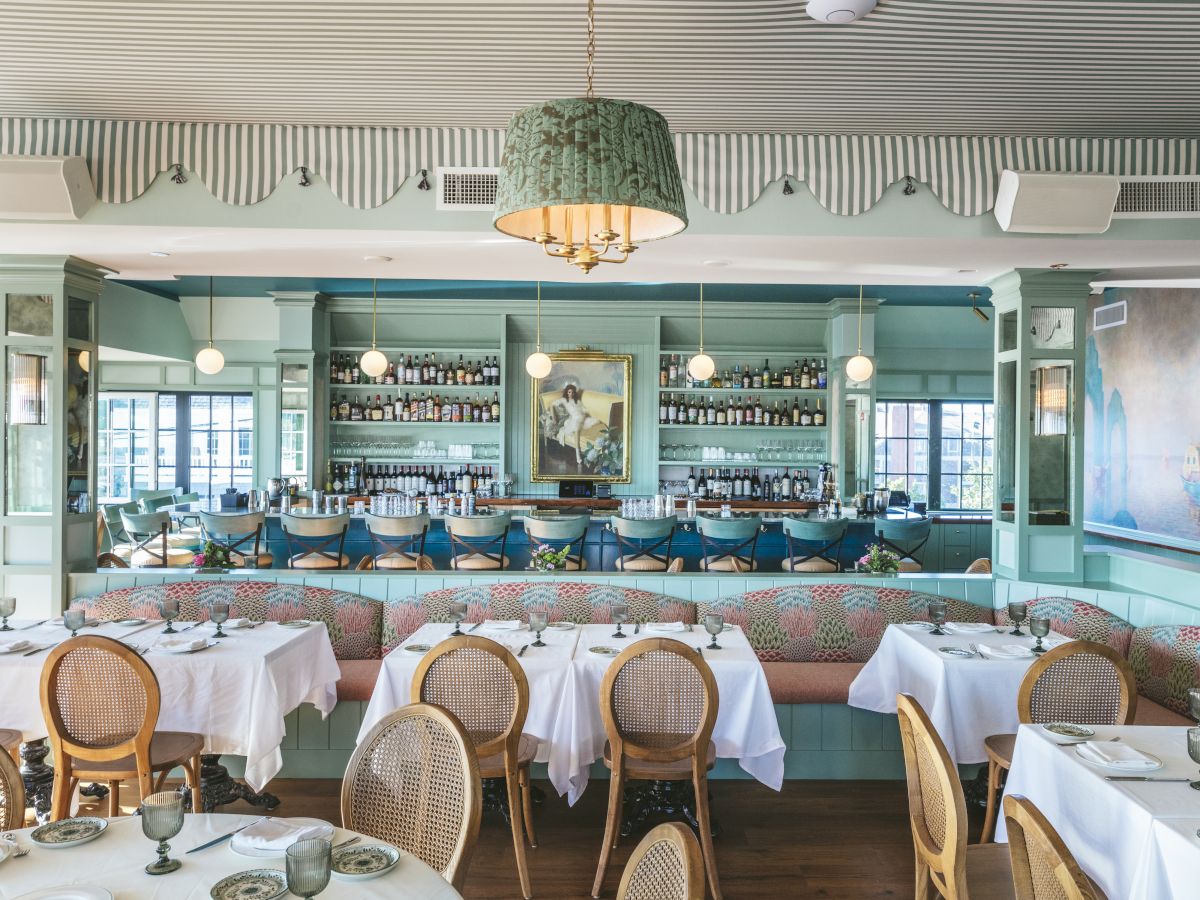 The image shows a stylish restaurant interior with neatly set tables, wicker chairs, a bar in the background, and elegant decor in soft pastel colors.