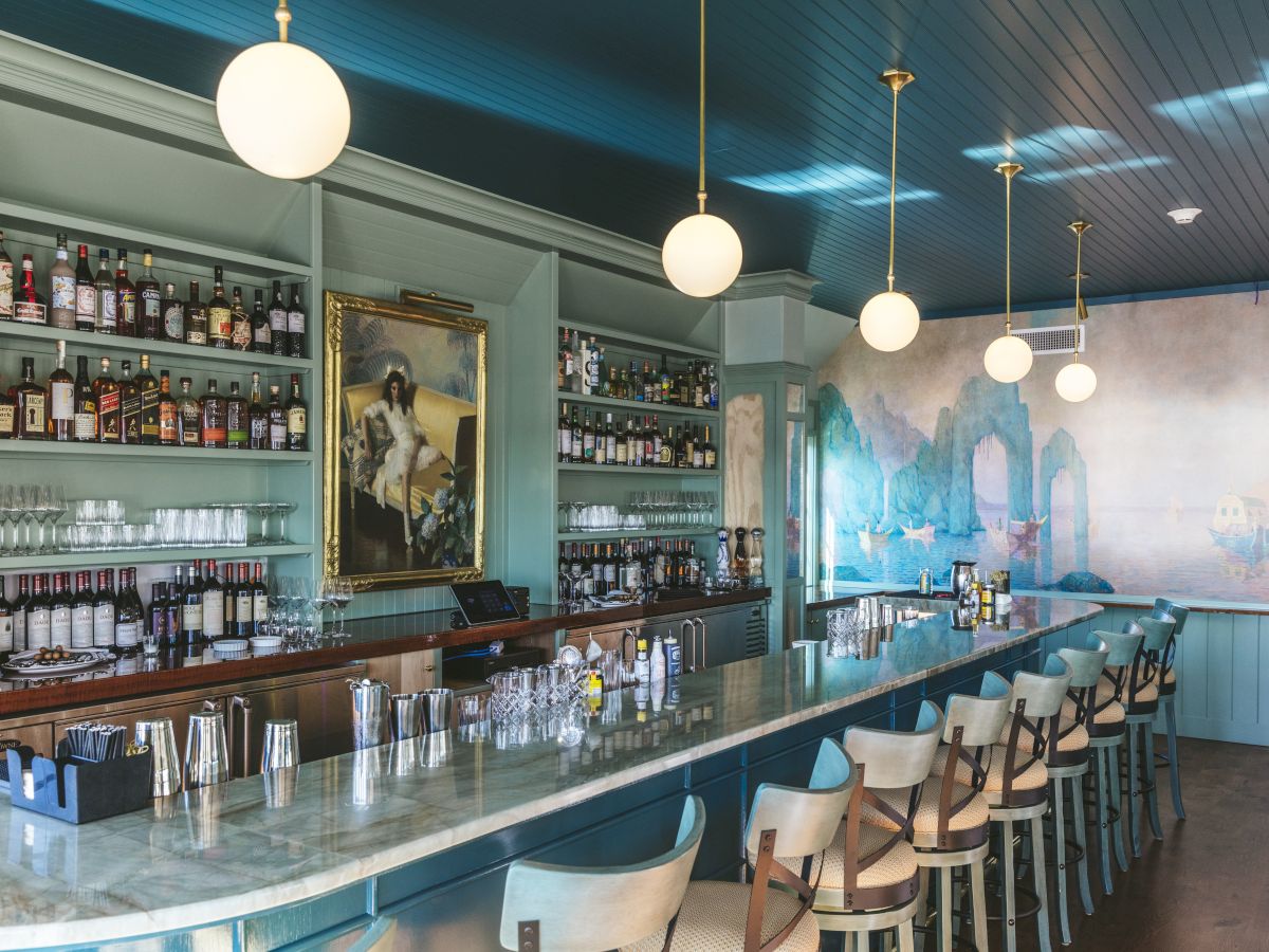 A stylish bar with a long counter, hanging lights, stocked shelves, a painting, and teal-themed decor with chairs lining the bar.