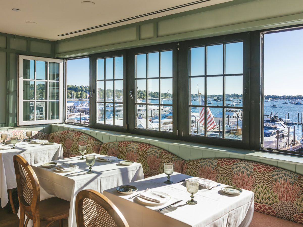 A cozy indoor dining area with wooden chairs and tables set, overlooking a scenic waterfront through large windows, perfect for a relaxed meal.