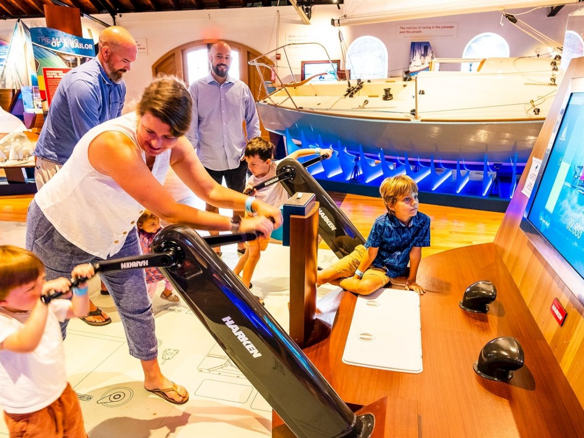 A woman and kids interact with a museum boat simulator, with staff nearby and a screen displaying a nautical scene.