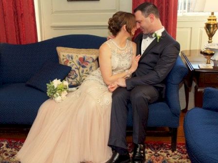 A wedding couple in formal attire share a tender kiss while seated on a blue sofa in a decorated room.
