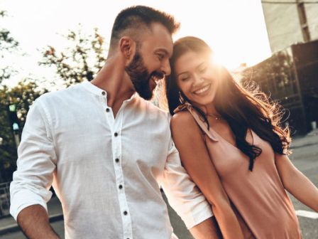 A smiling couple outdoors, the man in a white shirt and the woman in a brown dress leaning close in warm sunlight.