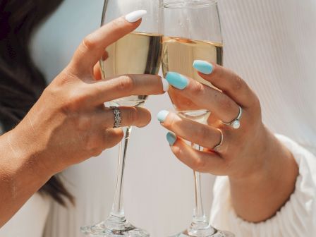 Two people clink champagne flutes in a toast, with pale sparkling drinks and pastel manicures visible.