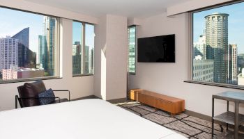 A modern hotel room with large windows showing a cityscape, a TV on the wall, a bed, a chair, and a small table.