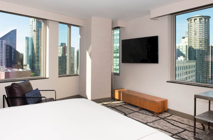 A modern hotel room with large windows showing a cityscape, a TV on the wall, a bed, a chair, and a small table.