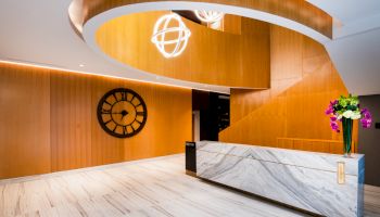 A modern lobby with a curved wooden staircase, a large wall clock, a marble reception desk adorned with flowers, and geometric light fixtures.