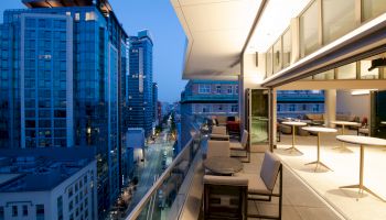 An urban balcony at dusk with outdoor seating overlooks a city street and tall buildings, featuring a modern design and a glass railing.