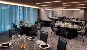 A modern banquet hall with round tables set for dinner, featuring elegant table settings, black napkins, and floral centerpieces under soft lighting.