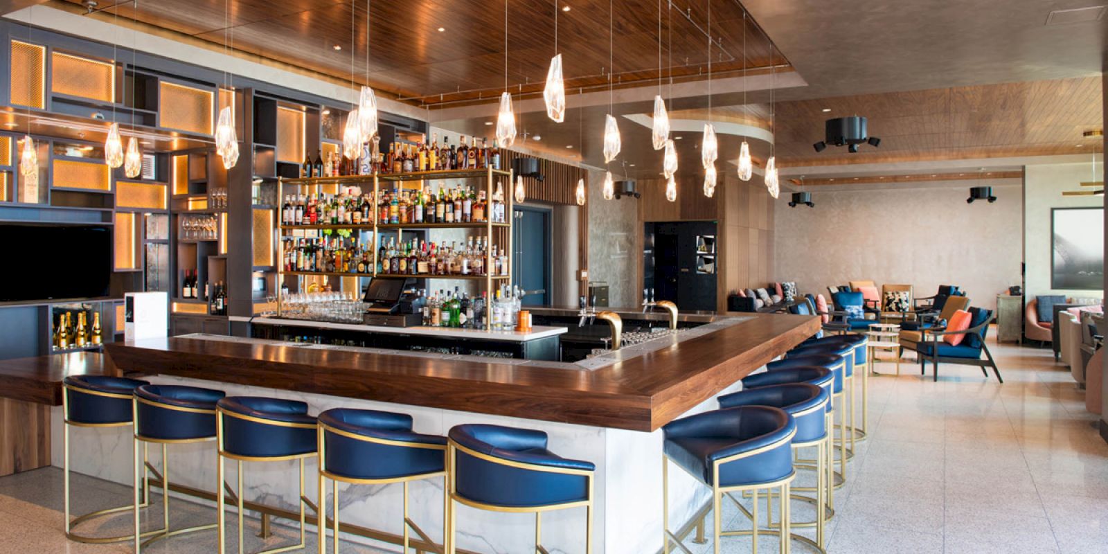 A modern bar area with high chairs, a well-stocked liquor shelf, and hanging pendant lights, located in a sleek, open space with additional seating.