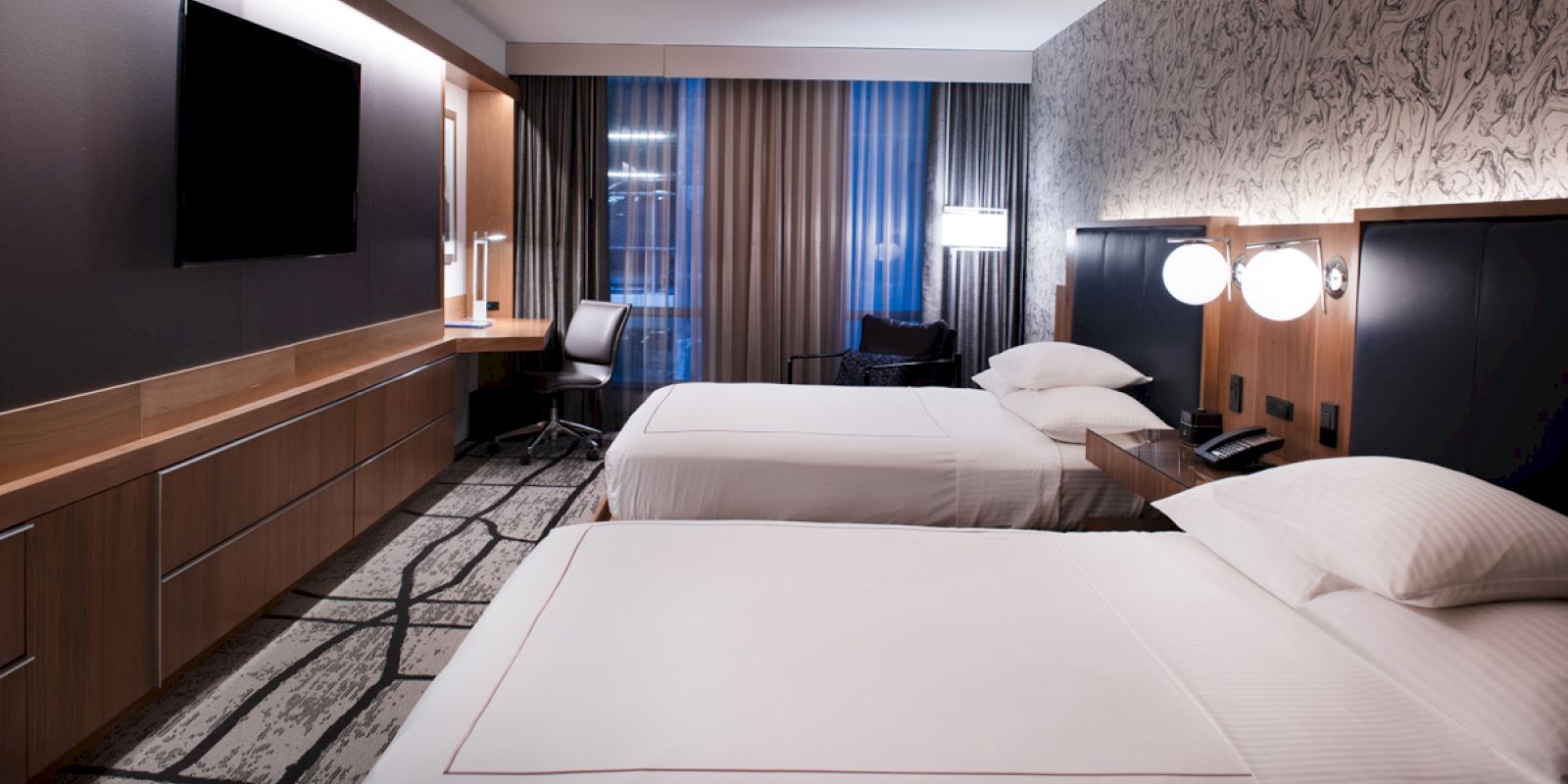 A modern hotel room with two double beds, a flat-screen TV, a desk with a chair beside the window, and stylish decor and lighting.