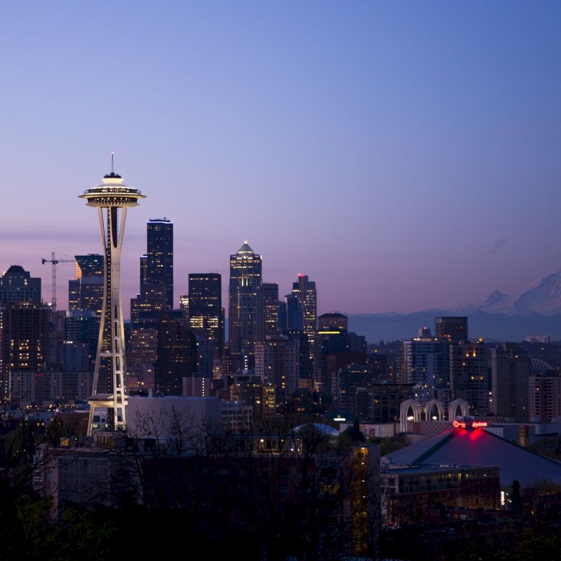 The image shows the Seattle skyline at dusk, featuring the Space Needle and downtown buildings with Mount Rainier in the background.