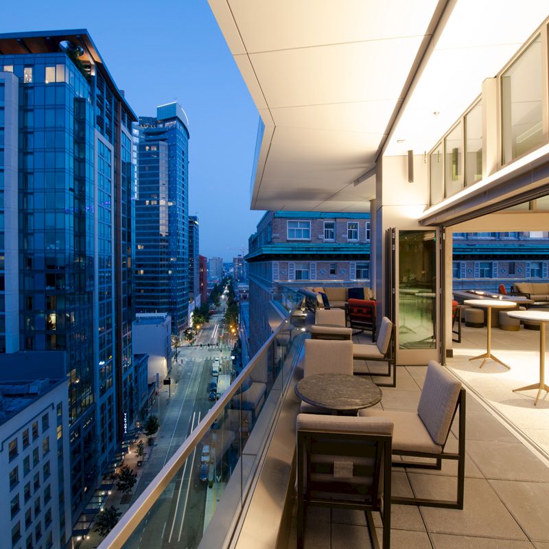 Image of an urban balcony at twilight, featuring a street view with tall buildings, outdoor seating areas with tables and chairs, and indoor lighting.