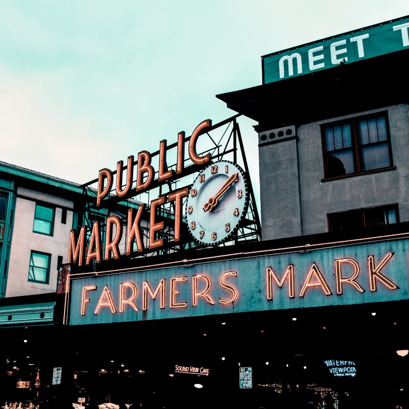 The image shows a public market with a large clock and a neon sign advertising a farmers market. There are buildings and signs in the background.