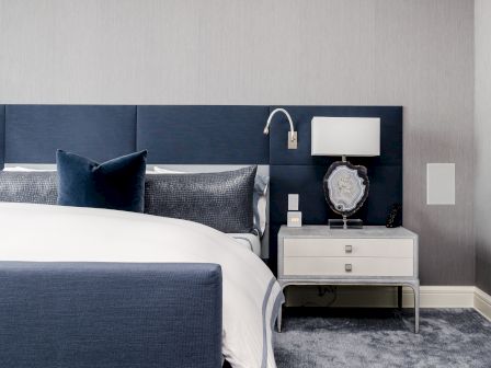 A modern bedroom with a navy blue upholstered headboard, a bed with white and blue bedding, and a bedside table holding a lamp and decorative object.