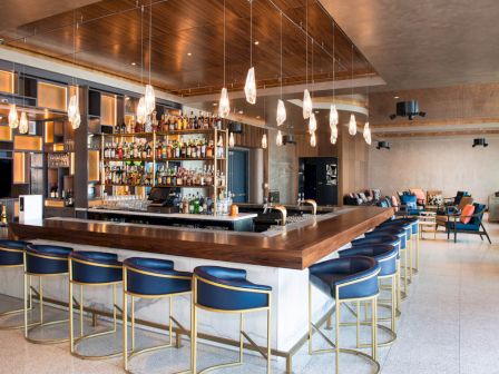 The image shows a modern bar area with blue bar stools, a wooden counter, hanging pendant lights, and shelves stocked with various bottles.