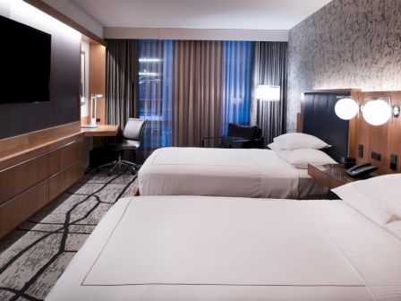 A modern hotel room with two beds, a TV, a desk, and a window with curtains. The decor features neutral tones and contemporary design elements.